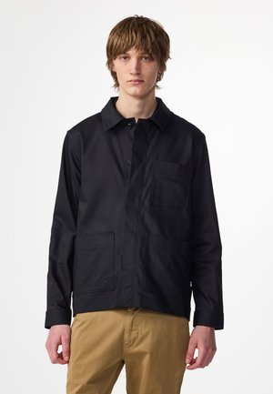 Giacca da lavoro nera con colletto, chiusura frontale a bottoni, due grandi tasche a toppa e un tessuto liscio e strutturato.