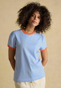 Frau mit lockigem Haar, die ein blau-weiß gestreiftes T-Shirt mit orangem Besatz trägt, steht vor einem schlichten beigen Hintergrund.