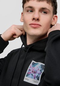 Sudadera negra con un bolsillo frontal, que presenta un gráfico floral colorido en un parche cuadrado. La tela parece suave y tiene un corte relajado.