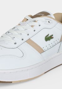 Λευκό και μπεζ sneaker Lacoste με λευκά κορδόνια και πράσινο λογότυπο κροκόδειλου στην πλευρά, εμφανίζεται σε κοντινή λήψη.