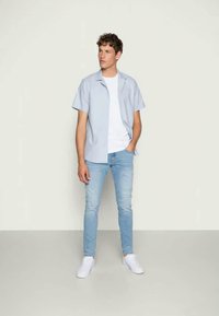 Hellblaues kurzärmliges Hemd über einem weißen T-Shirt, mit hellblauen Jeans und weißen Turnschuhen. Weicher Stoff mit lässiger Passform.