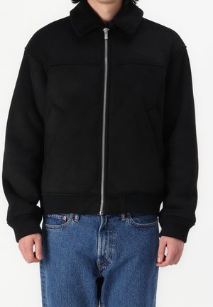 Homme portant une veste noire zippée avec col duveteux et jean bleu en denim, debout devant un fond blanc.