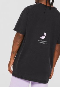Camiseta negra de gran tamaño con un gráfico de pluma rosa pastel en la parte posterior y texto blanco, hecha de tela de algodón para un ajuste informal.