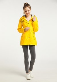 Gul vattentät jacka med dragkedja fram, grå huva, knappade ärmslut och sidofickor. Bärs över grå slim fit-jeans och vita sneakers.