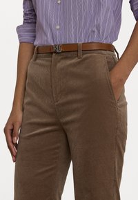 Pantaloni in corduroy marroni con righe verticali, abbinati a una camicia a righe viola chiaro. Una cintura in pelle marrone con una fibbia argentata con il logo "RL".