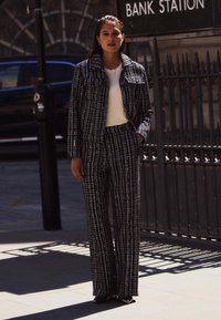 Svartvit tweedkostym med jacka och vida byxor, med texturerat tyg och måttanpassad passform. Modellen står mot en urban bakgrund.