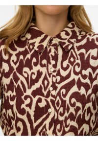 Chemise boutonnée à col avec un motif abstrait en spirale bordeaux et beige et un tissu texturé portée par une personne aux longs cheveux blonds.