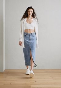 Frau, die nach vorne geht, trägt ein weißes Crop-Top, ein offenes weißes Hemd, einen blauen Jeansrock mitSchlitz vorne und weiße Sneaker auf einem Holzboden.