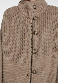 Beige-neuletakki, jossa on korkea kaulus, suuret napit ja teksturoidut kuviot. Edessä aukot ja teksturoidut viimeistelyt hihoissa.