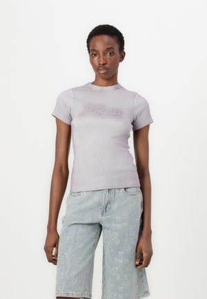 Istuva lilac t-paita lyhyillä hihoilla, ribbikuosisella pinnalla ja rintaneulalla, jossa on "Guess"-logo, yhdistettynä vaaleansinisisiin kulutettuihin shortseihin.