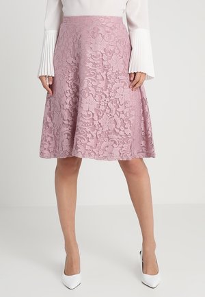 Femme portant une jupe trapèze en dentelle rose longueur genou, associée à un haut blanc à manches longues et des escarpins blancs pointus.