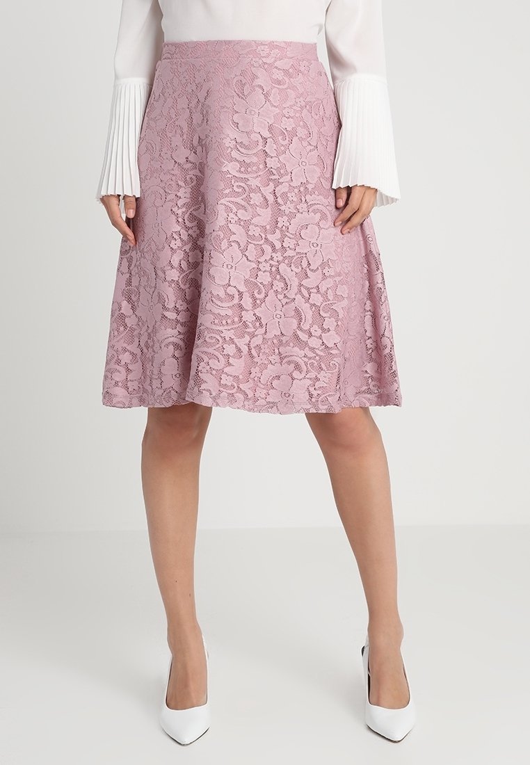Femme portant une jupe trapèze en dentelle rose longueur genou, associée à un haut blanc à manches longues et des escarpins blancs pointus.