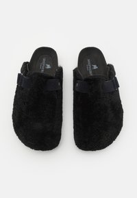 Črne, puhaste copate z mehko teksturo, okroglo obliko in tekstilnim trakom čez vrh. Zasnova za hitro obuvanje z oblazinjenim vložkom.