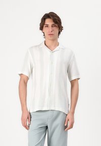 Camicia bianca a maniche corte, con bottoni e un motivo testurizzato e sottili righe pastello. Indossata con pantaloni azzurri chiari, caratterizzati da una vestibilità rilassata.