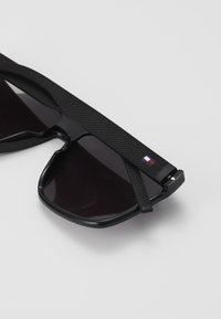 Lunettes de soleil noires avec une monture texturée noire, des verres sombres et un petit logo tricolore sur le côté. Le design présente une forme sportive et angulaire.