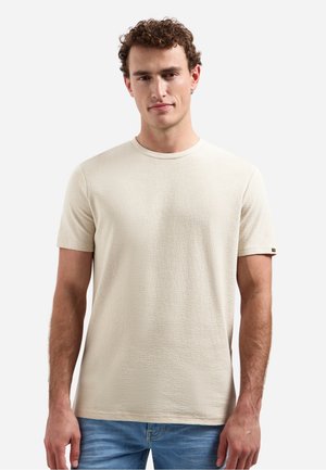 Man met krullend haar die een effen beige, gestructureerd t-shirt met korte mouwen en een blauwe spijkerbroek draagt, staand tegen een witte achtergrond.