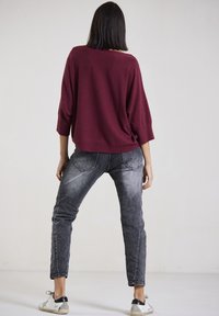 Burgunderfarbener oversized Pullover mit weiten Ärmeln, kombiniert mit enganliegenden grauen Jeans. Die Schuhe sind weiß mit schwarzen Akzenten. Rückansicht, glatte Textur.
