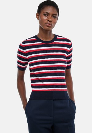 Femme debout, les mains dans les poches, portant un pull à manches courtes côtelé avec des rayures horizontales rouges, blanches et bleu marine, ainsi qu'un pantalon bleu marine.