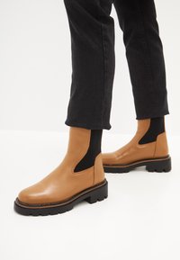 Botas de tobillo de cuero color marrón con una suela de goma negra gruesa, paneles elásticos laterales y detalle de costura a lo largo de la base. Llevadas con jeans negros.