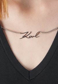 Juodas metalinis kaklo papuošalas su kursyvo stiliaus "Karl" pakabuku, prisiūtas prie sidabrinės grandinėlės; dėvimas prieš juodą V formos iškirptę.