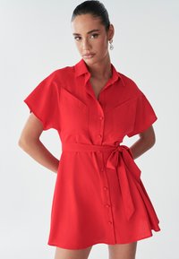 Model poseert zelfverzekerd in een korte, felrode jurk met een kraag en een strik in de taille, met knoopdetails en een zachte achtergrond.