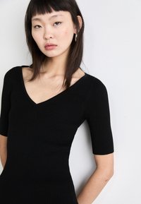 Črna rebrasta obleka z oprijetim silhuetom, kratkimi rokavi in V-izrezom, ki prikazuje gladko teksturo in strukturiran dizajn.