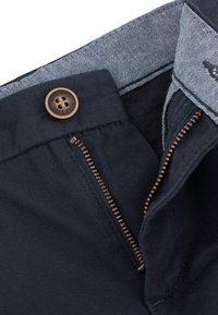 Gros plan d'un pantalon bleu marine avec une fermeture éclair métallique partiellement ouverte et un bouton marron portant l'inscription Authentic Next Dude.