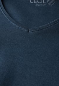 Námořnická modrá bavlněná tričko s výstřihem do V. Obsahuje jemné žebrování kolem výstřihu a hladkou texturu. Název značky viditelný v horní části.