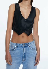 Schwarz-weiß gestreifte, bauchfreie Weste mit tiefem V-Ausschnitt, drei Knöpfen, tailliertem Schnitt und zwei Vorderentaschen. Kombiniert mit hochtailliertem Denim.