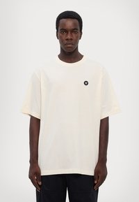 Beige oversize t-skjorte laget av myk bomull. Har rund hals og korte ermer, med en liten svart logo på venstre bryst.