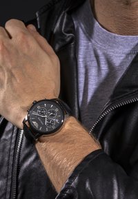 Melna hronogrāfa pulksteņa ar sudraba akcentiem, ādas siksna un apaļa sejas platne, kas parāda vairākas ciparnīcas un romiešu ciparus.