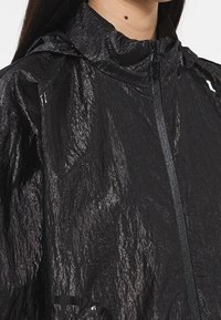 Kobieta ubrana w błyszczącą czarną, pomarszczoną kurtkę zapinaną na zamek z wysokim kołnierzem i kapturem, pokazująca górną część ciała i szyję.