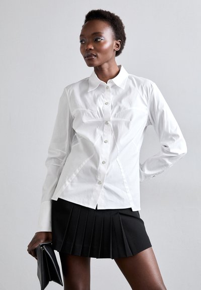 White button-up shirt with a fitted design and subtle seam detailing, paired with a black pleated skirt. Model holds a black clutch.
