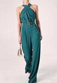 Mono halter en color verde azulado con cuerpo plisado y piernas anchas. Presenta una hebilla de cinturón redonda decorativa y se complementa con un pequeño bolso dorado.