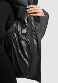 Melnā pūsto mētējs ar spīdīgu, guļamo oderi, ar rāvējslēdzēja kabatu un logo uzšuvi. Rokas pozīcija ir pie atvēruma.