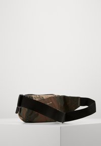 Kamuflážní ledvinka vyrobená z odolné tkaniny, disponující černým popruhem a zipovým uzavřením. Obdélníkový tvar s texturovaným povrchem.