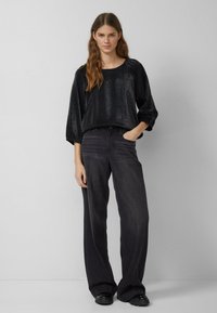 Schwarzes strukturiertes Oberteil mit lockerem Schnitt und ¾-Ärmeln, kombiniert mit hoch taillierten dunklen Denim-Jeans. Beide Kleidungsstücke sind einfarbig und haben eine glatte Oberfläche.