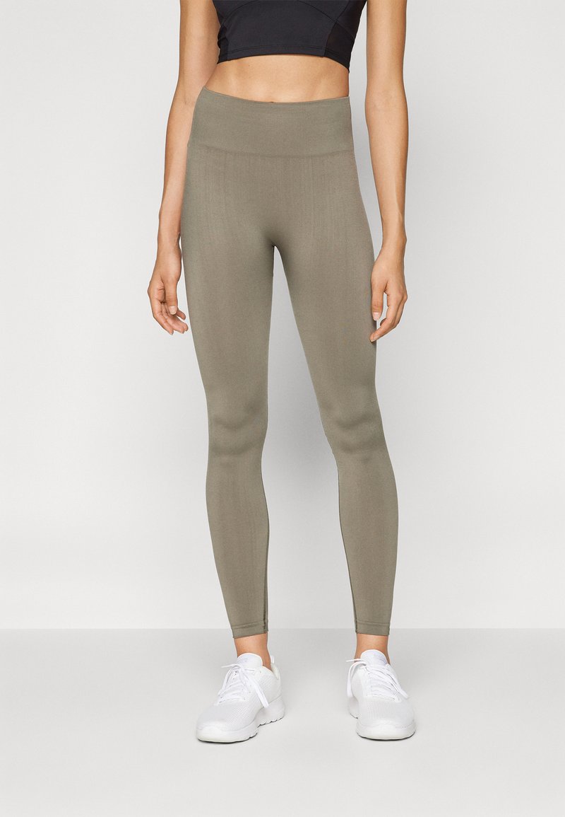 Des leggings vert olive en tissu extensible présentent une taille haute et une coupe slim, associés à des chaussures de sport blanches.