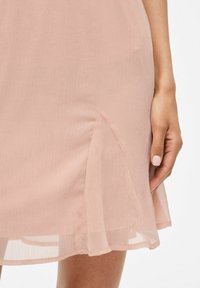 Abito rosa pallido realizzato in tessuto trasparente, con uno spacco laterale e una texture leggera. L'orlo è leggermente curvato. Mano visibile di lato.