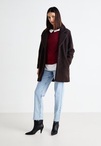 Manteau marron texturé avec de grands revers, porté sur un pull bordeaux et une chemise blanche, associé à un jean bleu clair et des bottes à talons noires.