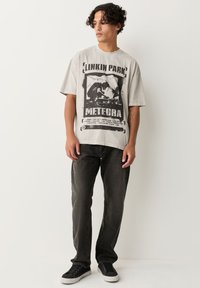 Giovane uomo che indossa una maglietta grigia dei Linkin Park Meteora, jeans neri e scarpe da ginnastica nere, in piedi contro un muro bianco.