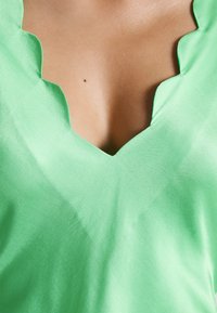 Grünes Satin-Top mit V-Ausschnitt und gewellten Kanten, das eine glatte Textur und eine lebendige Farbnuance präsentiert.