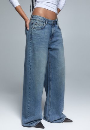 Kvinde iført blå jeans med høj talje og vide ben, hvid kortærmet langærmet top og sorte spidse sko, hånd i lommen.