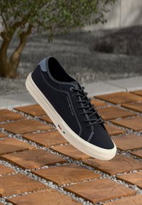 Baskets noires avec des accents gris, lacets plats et tige en tissu texturé. Semelle en caoutchouc blanche avec un petit logo près du talon.