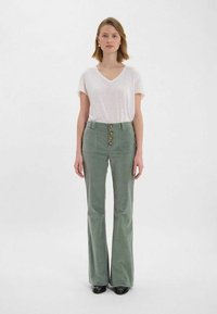 Pantalons évasés vert clair avec poches avant et fermeture à cinq boutons, associés à un t-shirt blanc à col en V ample. Texture lisse.
