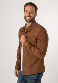 Bruine suède jas met een kraag halslijn, een voorzak en knoopsluiting. De stof heeft een zachte textuur en subtiele stikdetails.
