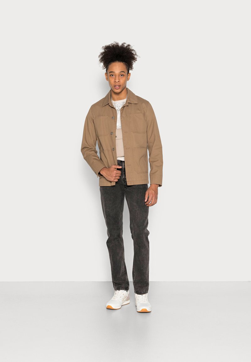 Junger Mann mit lockigem Haar, der eine beige Jacke, ein helles Hemd, dunkle Jeans und weiße Sneakers trägt. Er steht vor einer hellen, einfarbigen Wand.