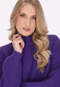 Fioletowy sweter z prążkowanym wzorem, z wysokim kołnierzem i długimi rękawami. Modelka delikatnie trzyma podbródek, podkreślając teksturowany splot swetra.