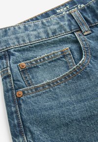 Jeans de denim con un lavado azul, con costuras en contraste, un bolsillo frontal con remaches y una etiqueta en la cinturilla.