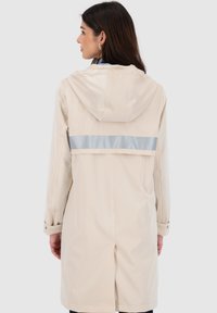 Impermeabile beige con cappuccio, caratterizzato da una striscia orizzontale lucida argento sulla schiena e maniche con bottoni. Texture liscia.